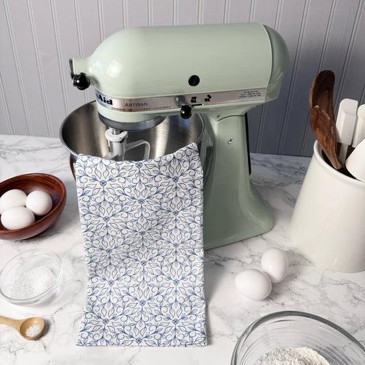 Light green KitchenAid mixer on a kitchen counter with eggs, flour, and a patterned towel.