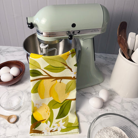 Kitchen scene with a green stand mixer, lemon-themed towel, and baking ingredients on a marble countertop.