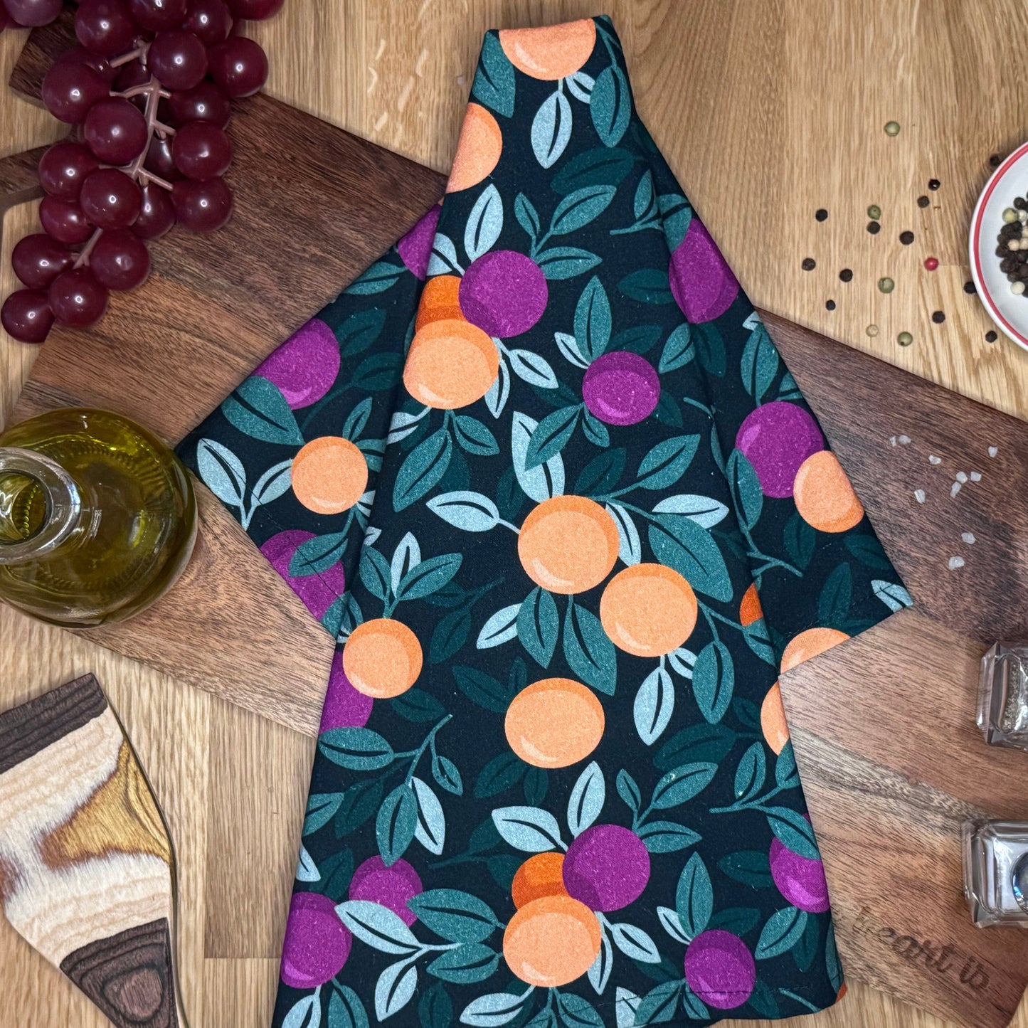 A multicolored tea towel with a pattern of oranges and purple berries displayed on a wooden surface, with some grapes and kitchen items in the background.