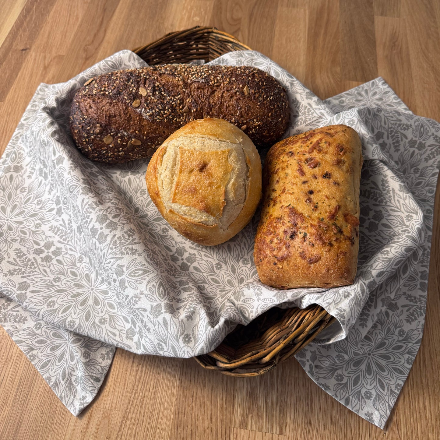Three loaves of bread in a basket on a wooden surface with a patterned cloth.