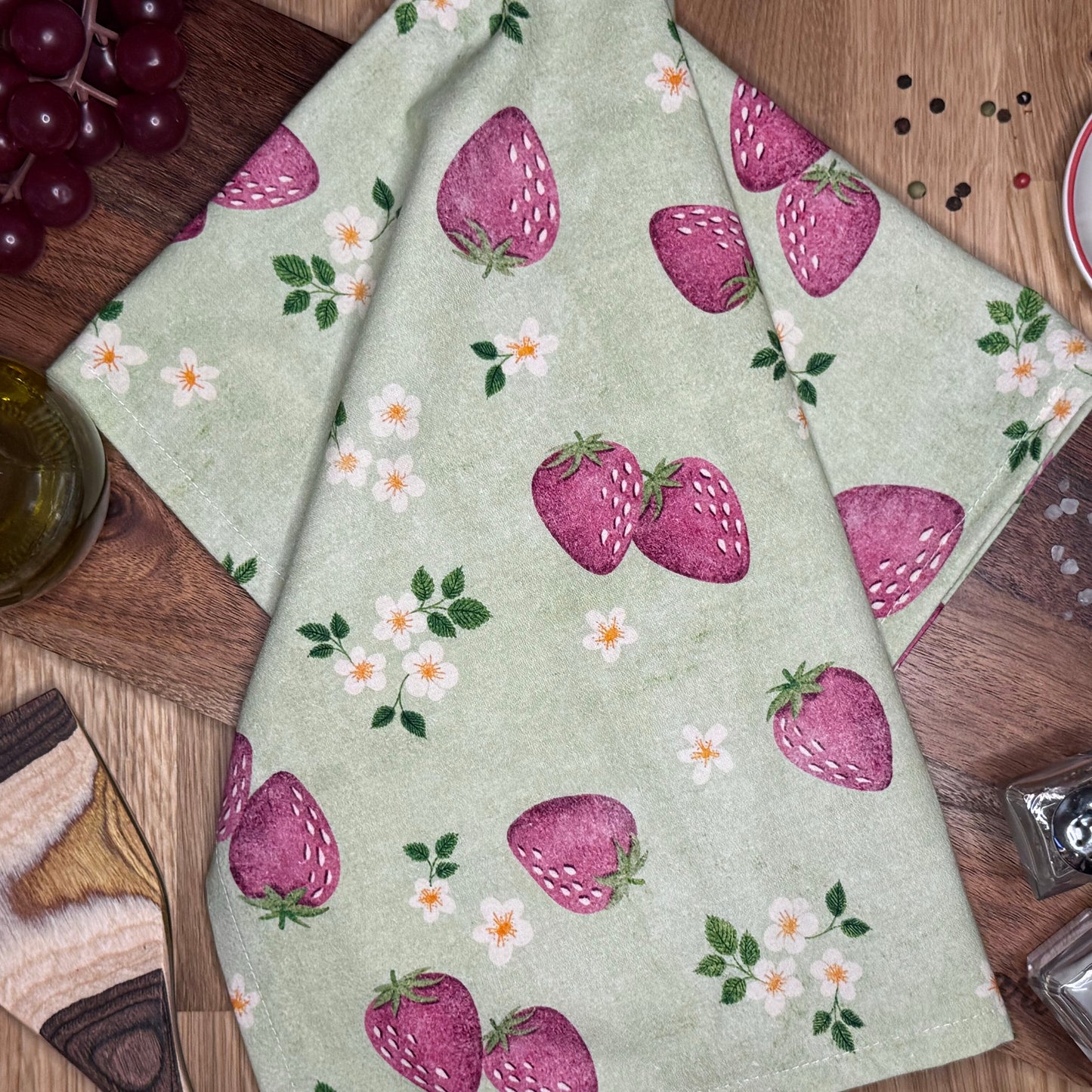 A green tea towel with a hand-illustrated pattern of red strawberries and white flowers.