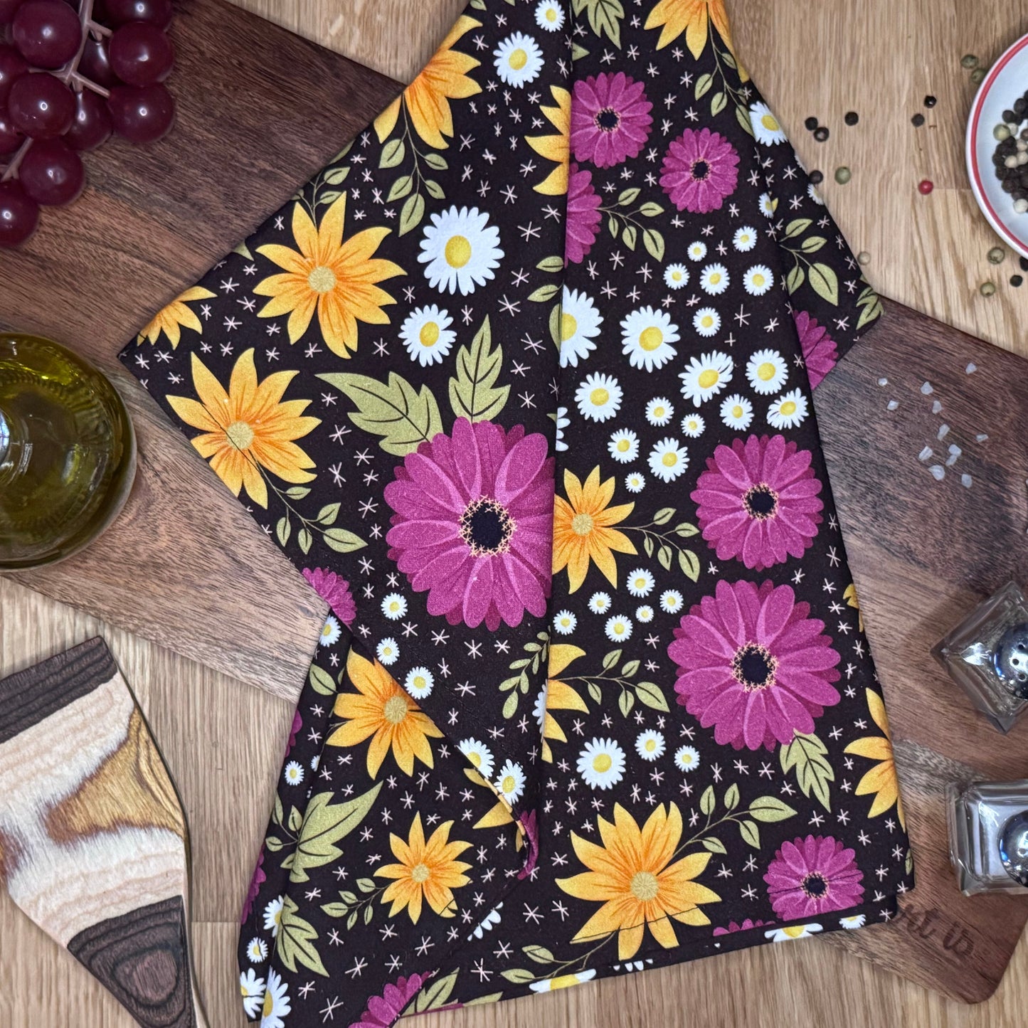 A multicolored floral tea towel with prominent sunflower and daisy designs, displayed on a wooden surface.
