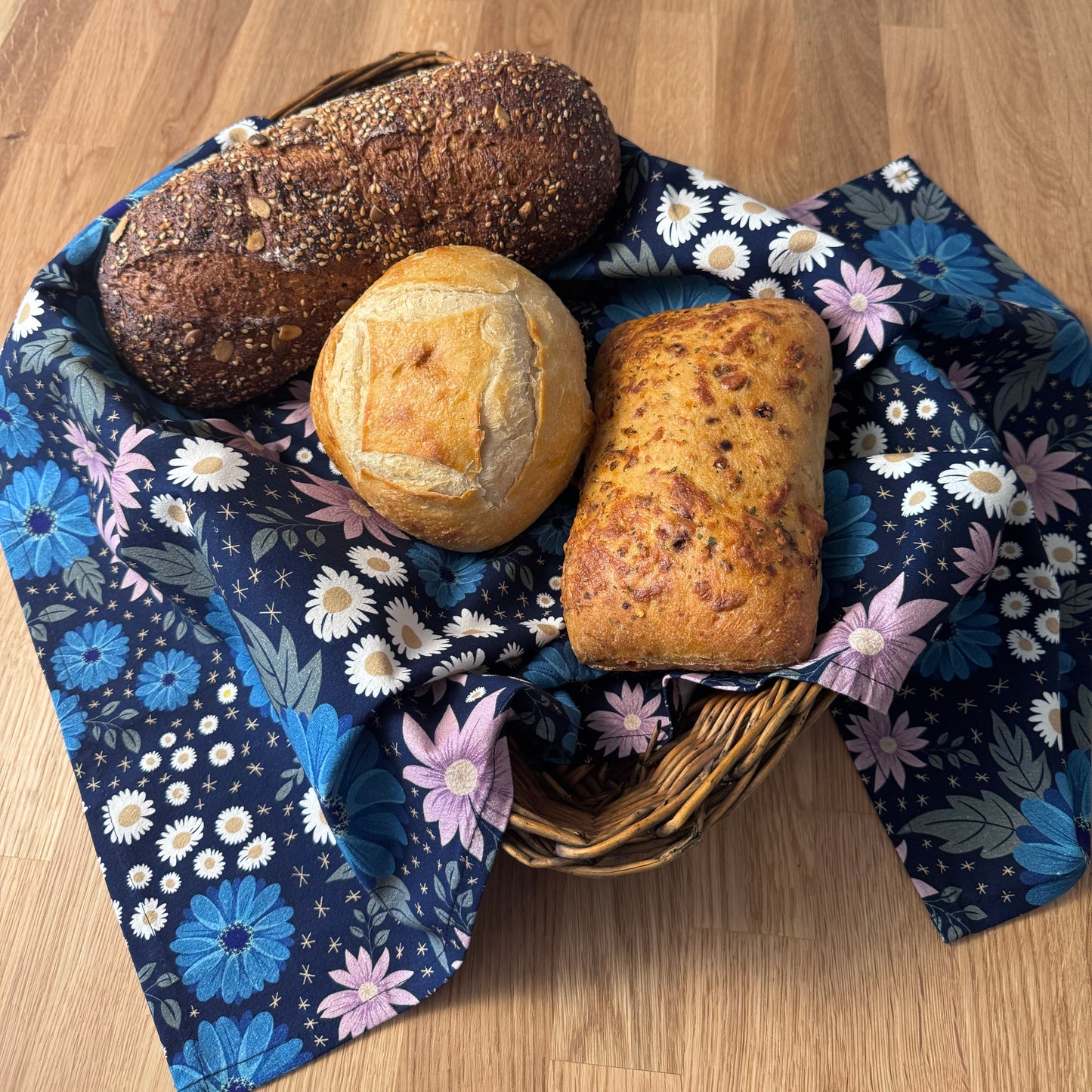 Three loaves of bread in a basket with a floral cloth on a wooden surface