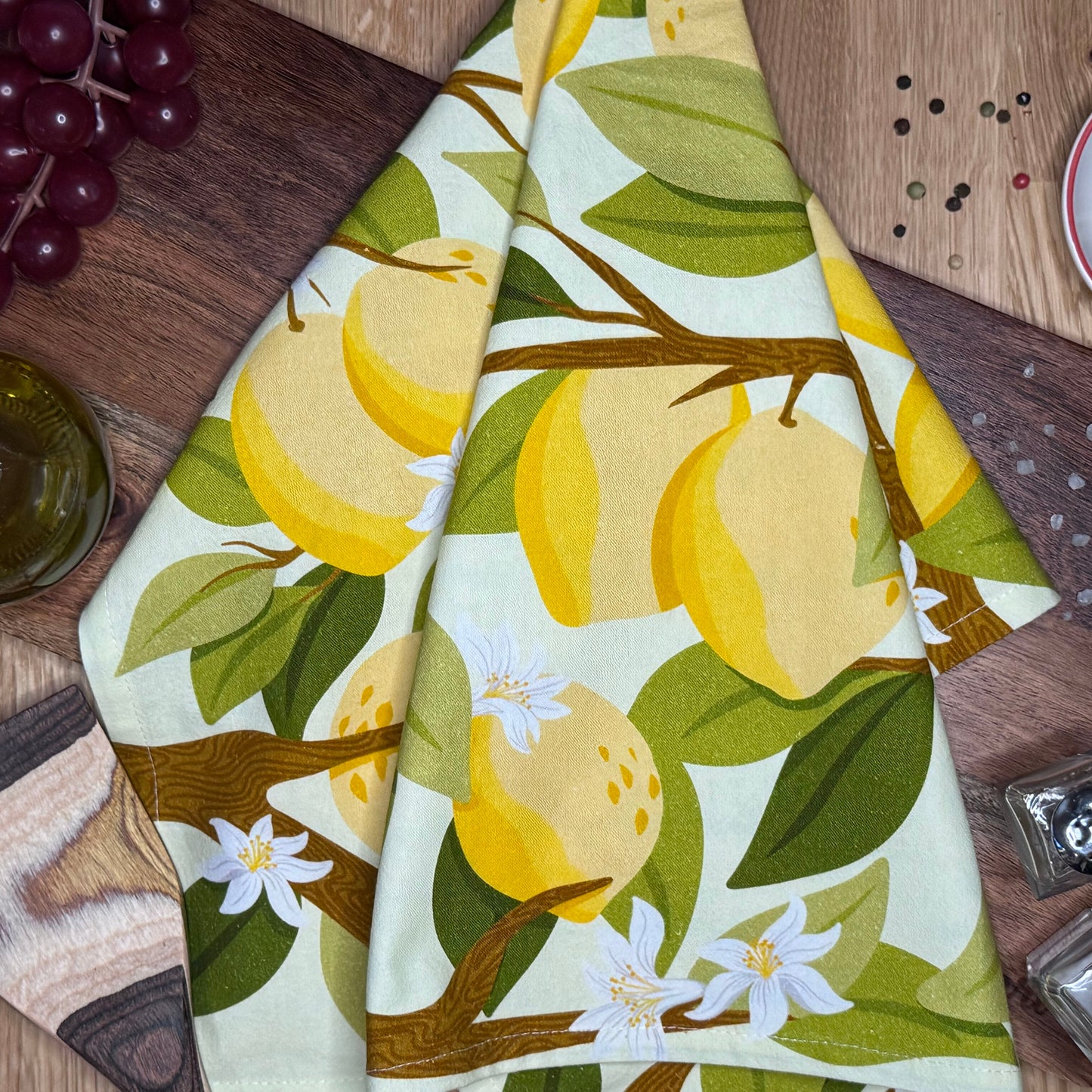 A folded cotton tea towel with a botanical lemon and leaf print, laid out on a wooden surface.