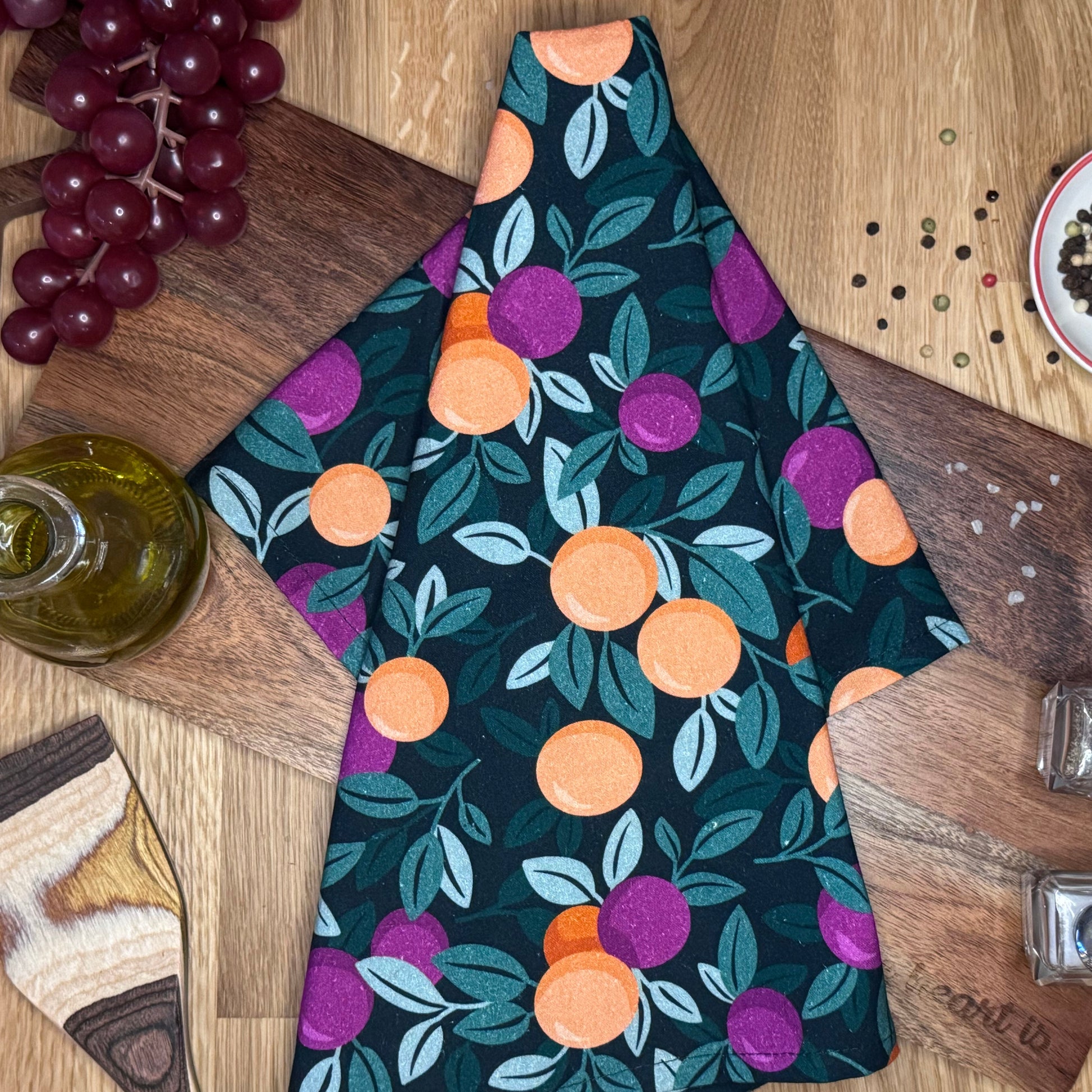 A multicolored tea towel with a pattern of oranges and purple berries displayed on a wooden surface, with some grapes and kitchen items in the background.