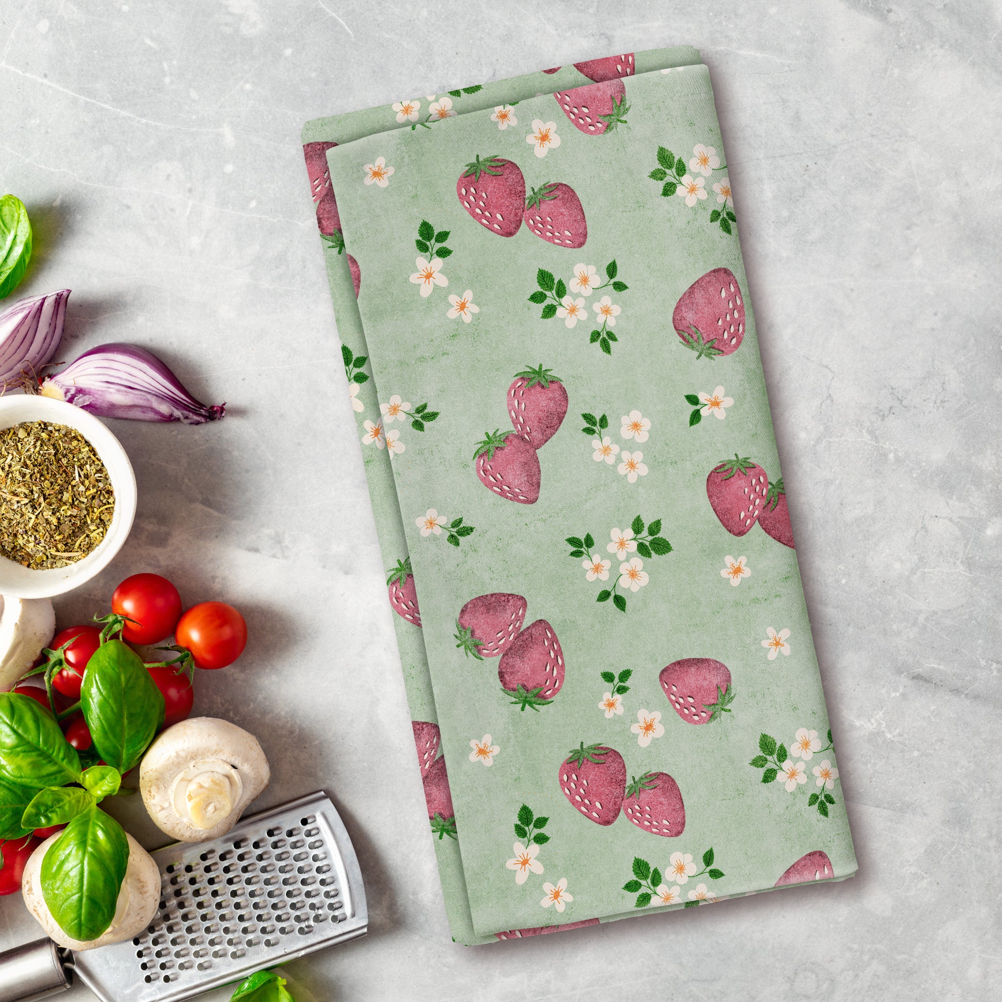Green kitchen towel with strawberry pattern on a gray surface with vegetables and a grater.