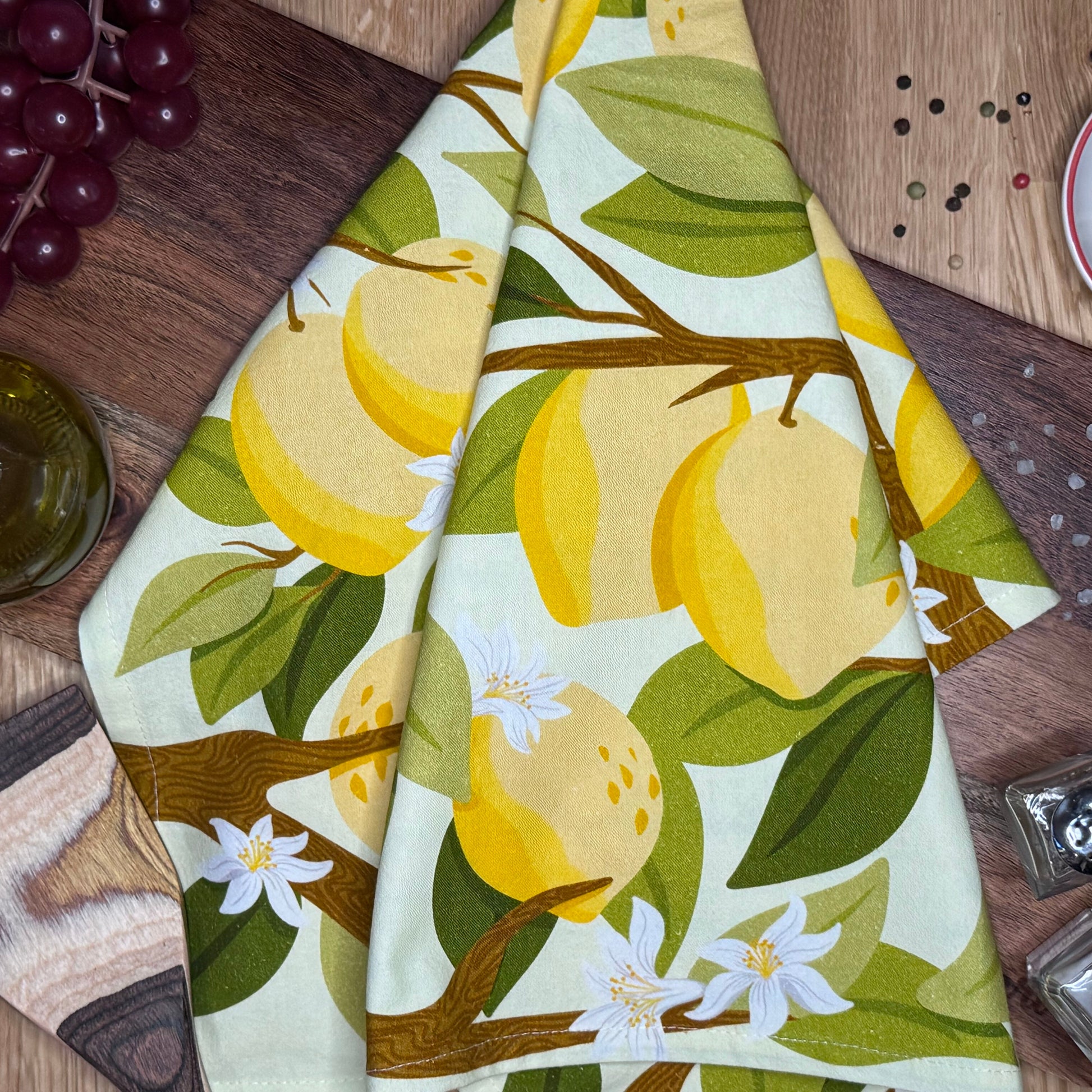 A folded cotton tea towel with a botanical lemon and leaf print, laid out on a wooden surface.