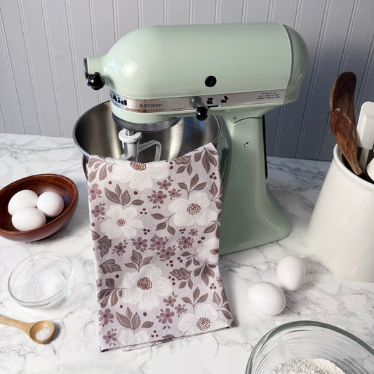 Mint green stand mixer on a kitchen counter with a floral towel, eggs, and kitchen utensils.